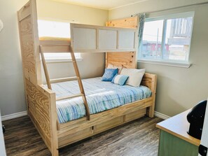 A wooden bunk bed with a ladder, a window with curtains, and a desk with a black object on it.