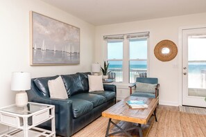 A living room with a dark leather sofa, a coffee table, a white side table, and a painting of sailboats. - Beach Barn - Nice 5BR Oceanfront Home in Heart of Surf City (Surf City)