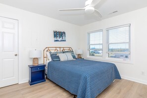 Ein Schlafzimmer mit einem Bett, einem Nachttisch, einem Deckenventilator, einem Fenster mit Jalousien und einem Wandbild.