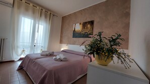 Une chambre à coucher avec un lit, une table de chevet ornée d’un vase de fleurs, un tableau au mur et une fenêtre avec des rideaux.