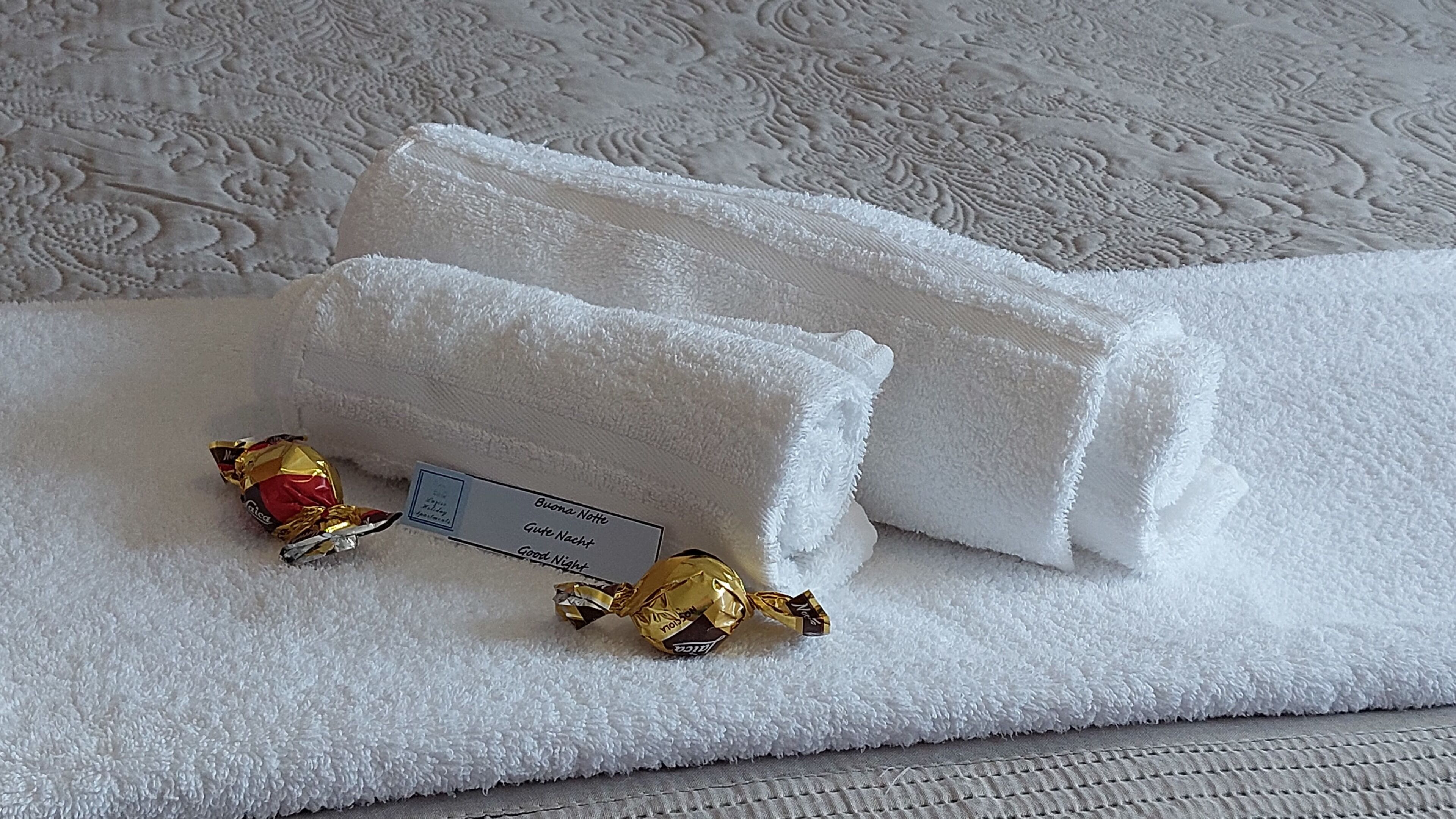 Une serviette blanche, soigneusement pliée, accompagnée d ’une petite carte et de deux chocolats emballés dans du papier doré, le tout posé sur une surface texturée.