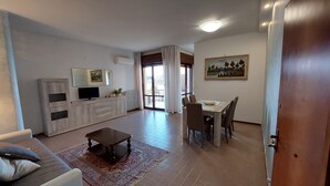 Un salon comprenant un canapé, une table basse, un téléviseur posé sur un meuble en bois, une table à manger avec des chaises et un tableau accroché au mur.