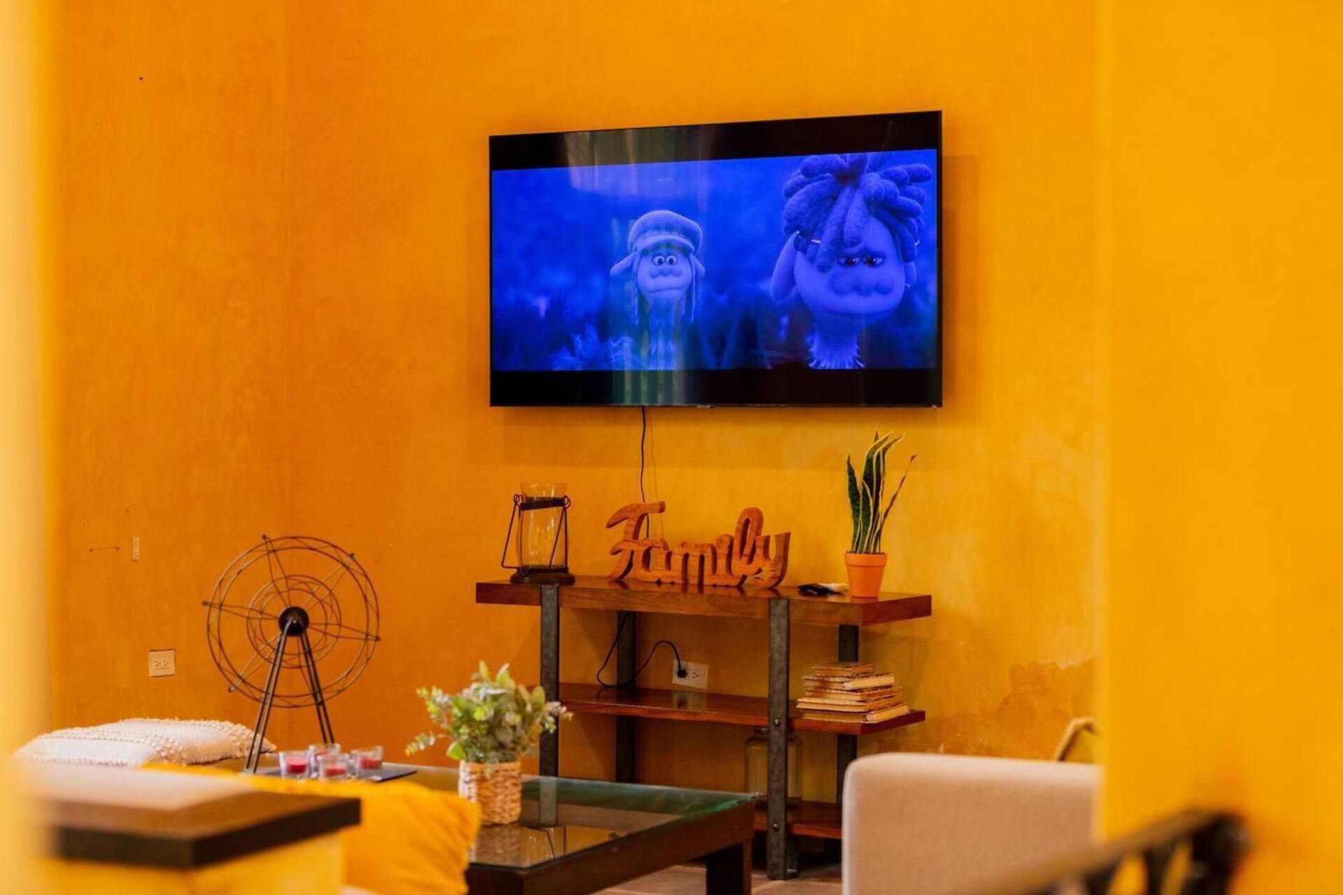 Una sala de estar con un televisor montado en la pared, un estante de madera con un letrero que dice 'Familia', una mesita con una planta y un ventilador en el suelo.