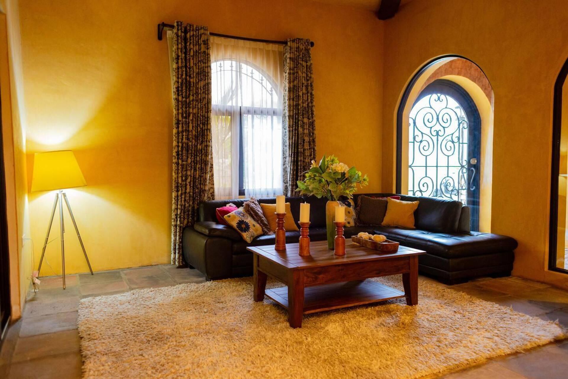 Una sala de estar acogedora con un sofá marrón oscuro, una mesa de centro de madera con velas y un amplio ventanal con cortinas.