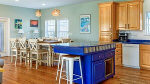 A kitchen with wooden cabinets, a blue island, and a dining area with white chairs. - Coconut Breeze - 105 St Dennis 4 Bedroom Home by RedAwning (Dauphin Island)