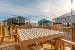 A wooden deck with a table and chairs, overlooking a beach with two houses and a clear sky. - Coconut Breeze - 105 St Dennis 4 Bedroom Home by RedAwning (Dauphin Island)