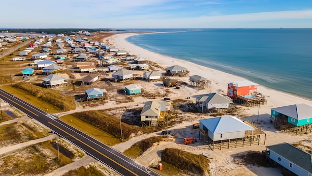 An aerial view of a coastal area with multiple buildings, some elevated on stilts, and a road running parallel to the beach. - Coconut Breeze - 105 St Dennis 4 Bedroom Home by RedAwning (Dauphin Island)