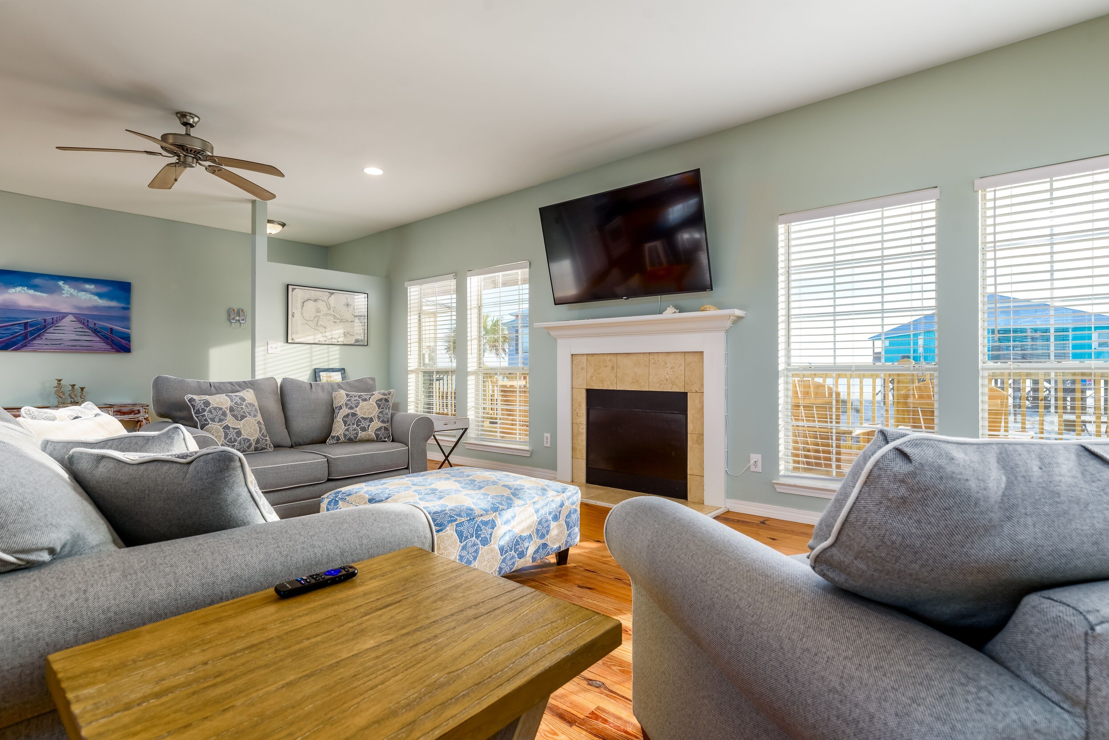 A living room with a fireplace, a ceiling fan, a TV, and a large window with blinds.