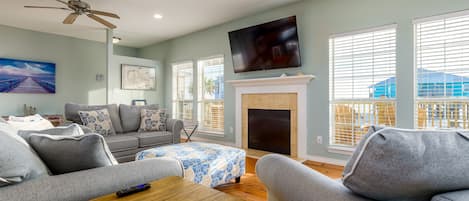 A living room with a fireplace, a ceiling fan, a TV, and a large window with blinds.