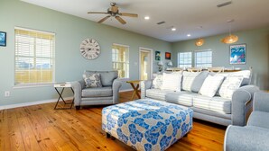 A spacious living room with a grey sofa, a blue and white ottoman, and a ceiling fan. - Coconut Breeze - 105 St Dennis 4 Bedroom Home by RedAwning (Dauphin Island)