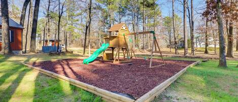 Un parque infantil con tobogán y columpios, rodeado de árboles y una casita de juegos de madera.