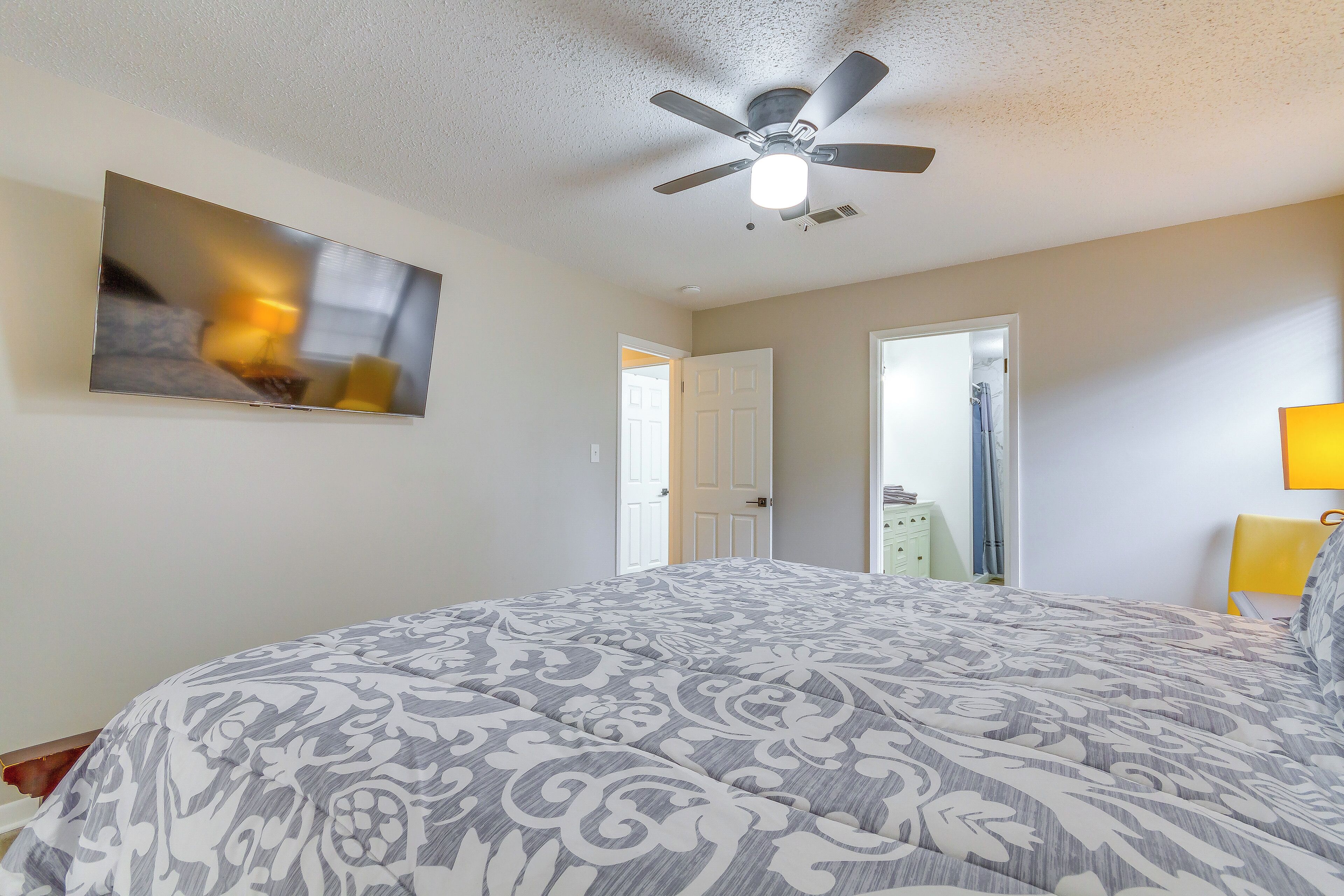 A bedroom with a bed, a ceiling fan, a TV mounted on the wall, and a door leading to another room.