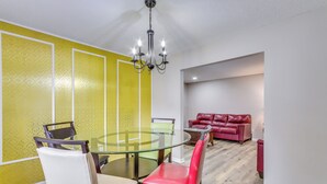 A dining area with a glass table and chairs, a chandelier, and a red leather sofa in the background. - Pensacola Home w/ Private Yard: 7 Mi to Downtown! (Pensacola)