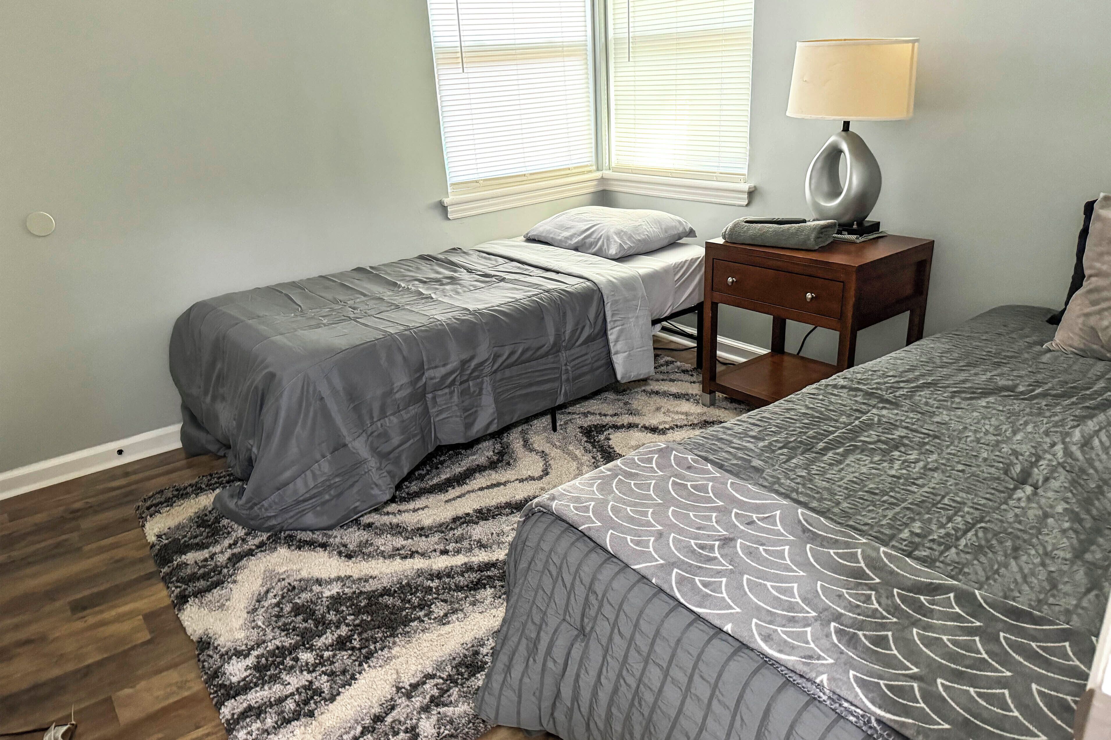 A bedroom with two beds, a nightstand with a lamp, a rug, and a window with blinds.