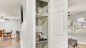Une pièce avec un plancher en bois, un coin repas avec une table et des chaises, un lit près d’une fenêtre et une armoire avec des étagères.