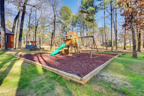 Une aire de jeux avec un toboggan et des balançoires, entourée d’arbres et agrémentée d’une cabane en bois.
