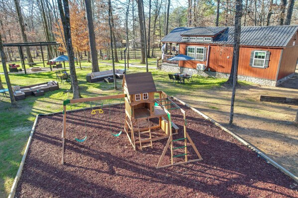 A playground with a wooden swing set and climbing structure, surrounded by a grassy area with benches and a small house-like building. - Hook, Line, & Sinker Retreat: Fire Pit, Grill! (Broken Bow)