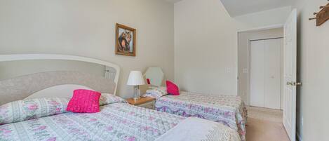 Une chambre avec deux lits, chacun agrémenté de draps à motifs floraux et d’un oreiller rose. On y trouve une table de chevet avec une lampe et un tableau encadré accroché au mur.