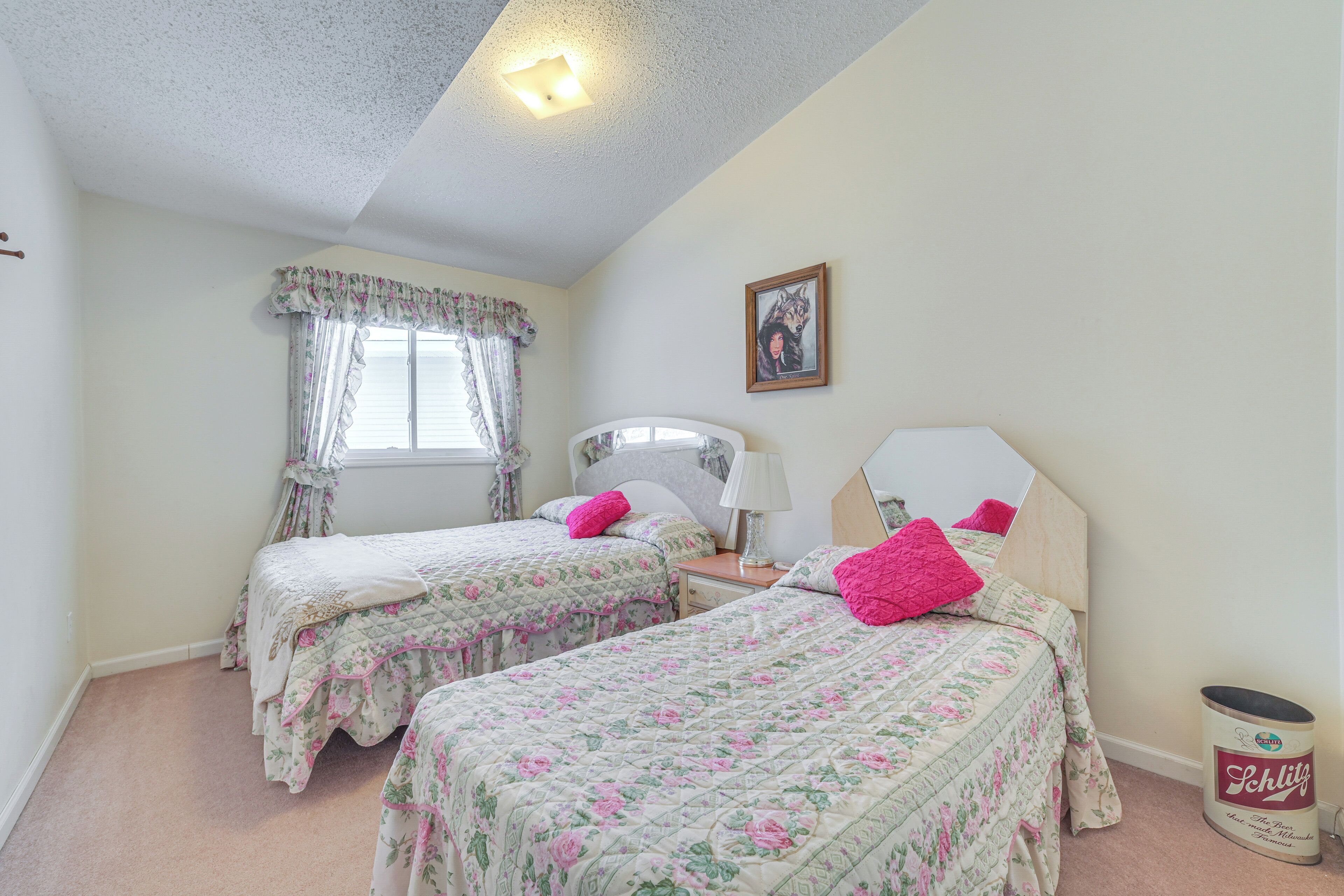 A bedroom with two beds, floral bedding, a window with curtains, a bedside lamp, and a picture on the wall.