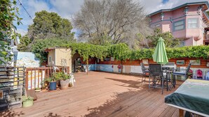 Eine Holzterrasse mit Gartenmöbeln, Blumentöpfen und einem Gebäude mit rosafarbenem Mauerwerk im Hintergrund.