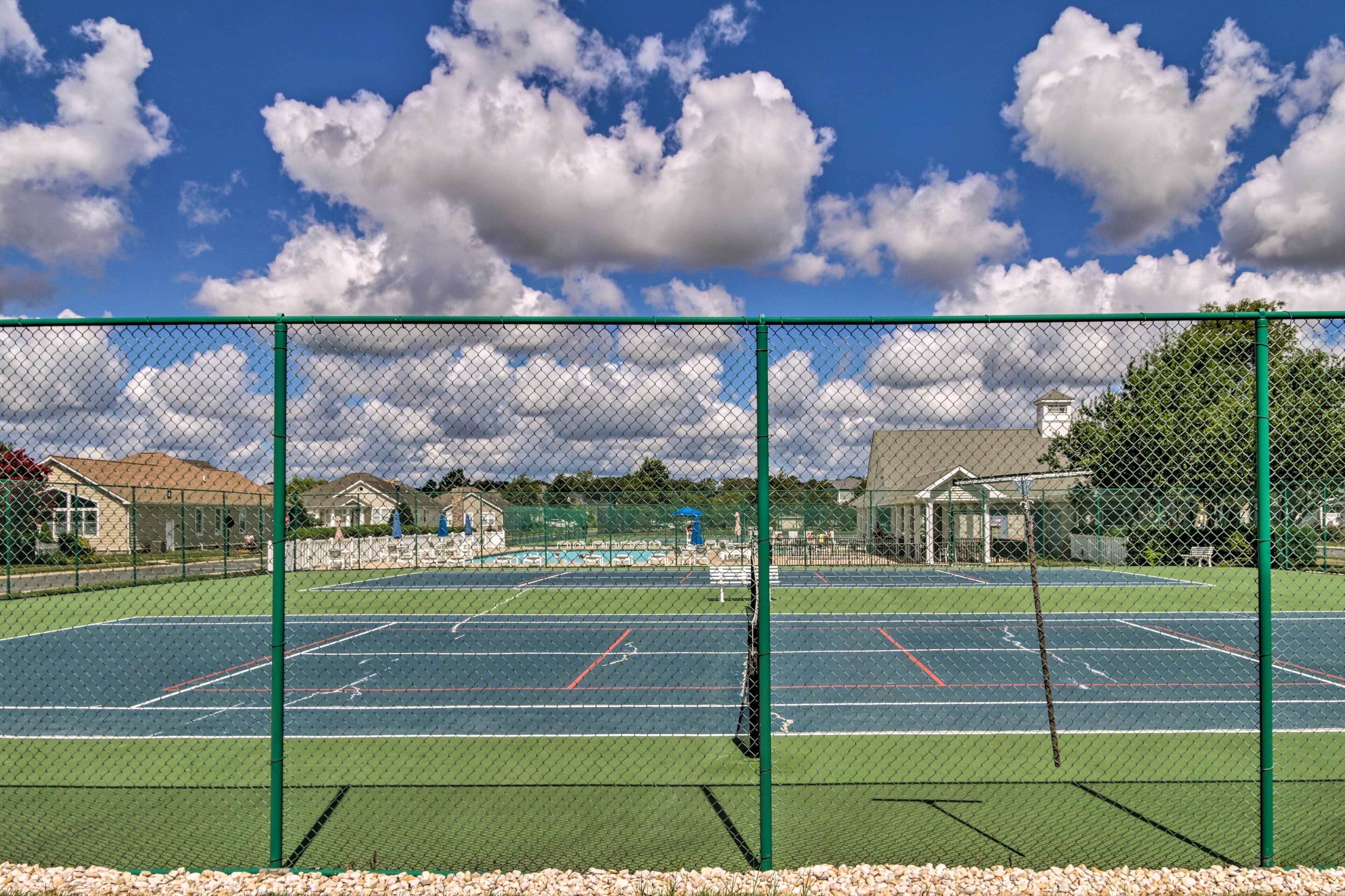 A tennis court with a green fence, surrounded by buildings and trees.