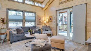 A cozy cabin interior with a gray sofa, a small round table, and a beige ottoman. There is a large window with blinds, a door leading outside, and a wooden ceiling. - 'sweet Spot' Cabin in Broken Bow w/ Fire Pit! (Broken Bow)