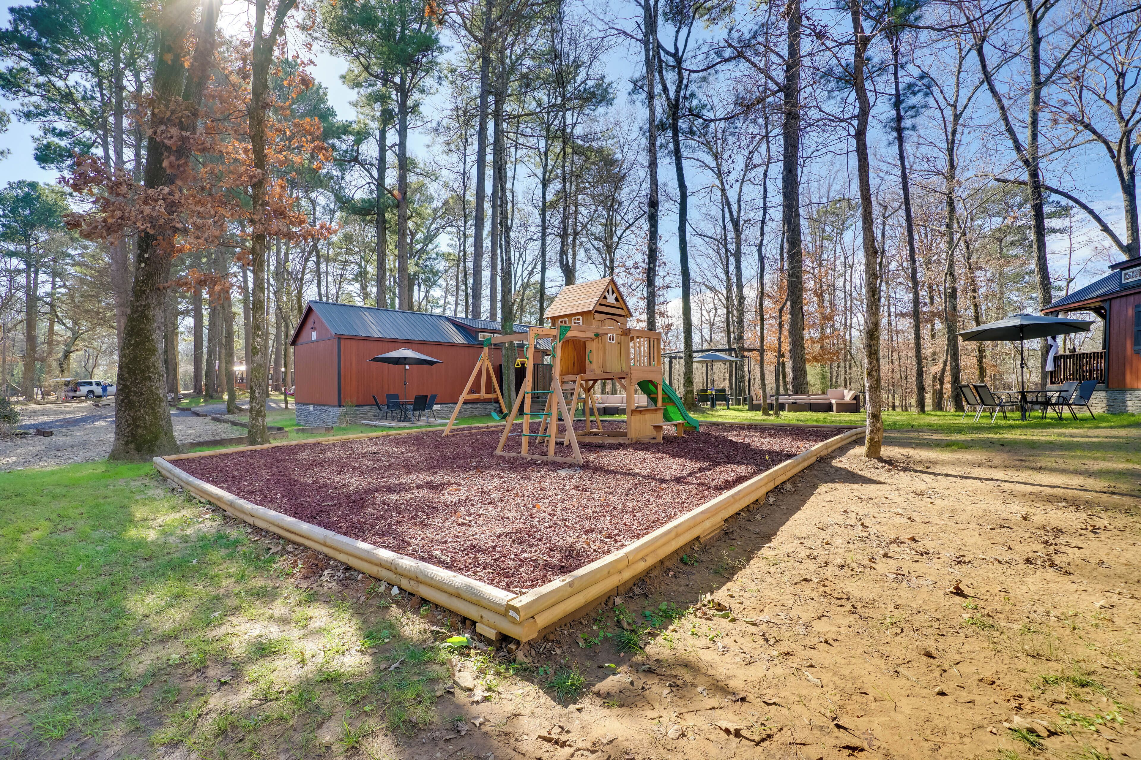A playground with a wooden playhouse, swings, and a slide, surrounded by trees and a dirt path.