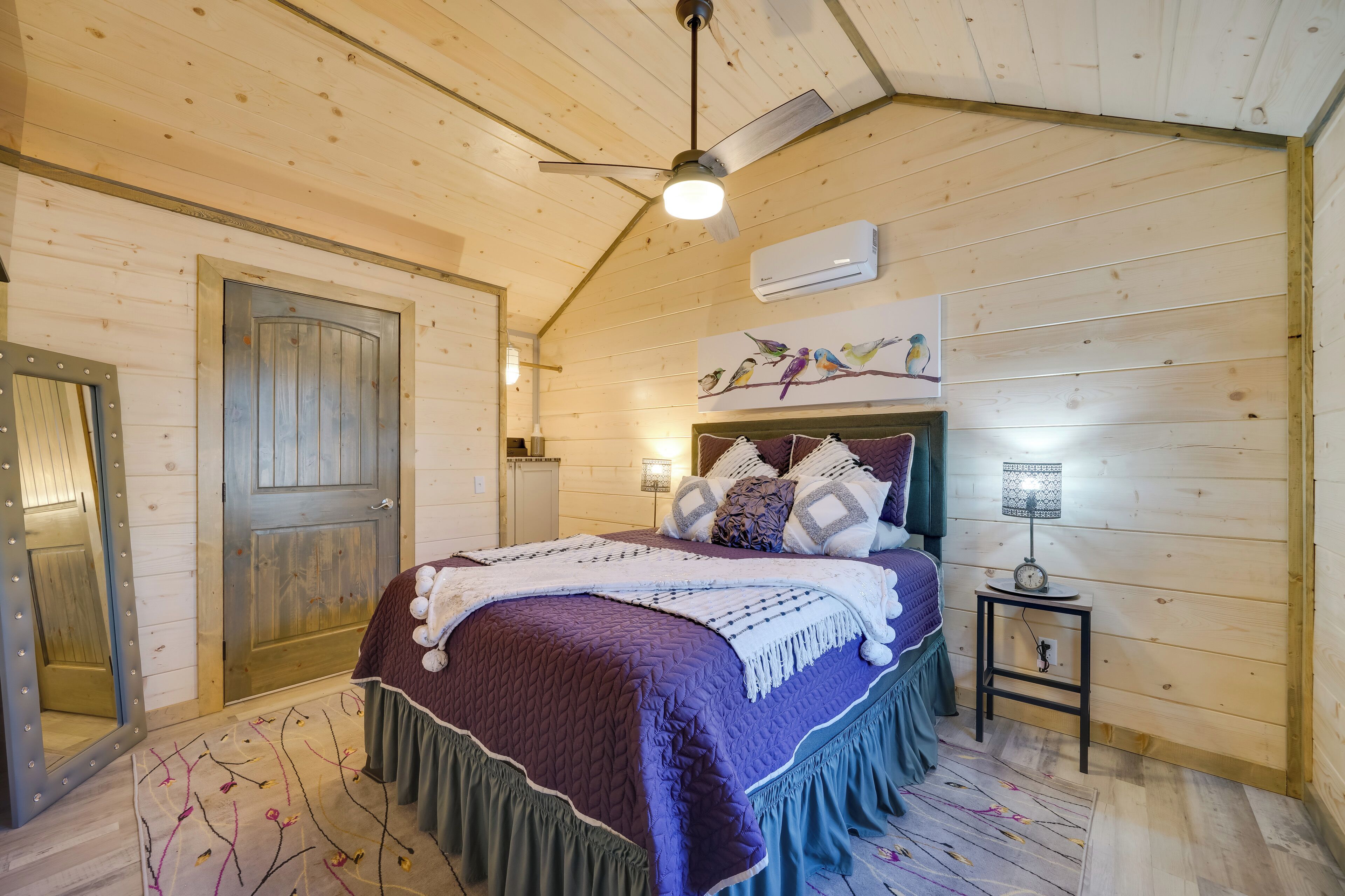 A cozy bedroom with a wooden ceiling, a bed with a purple and green bedspread, a small bedside table with a lamp, and a wall-mounted air conditioner.