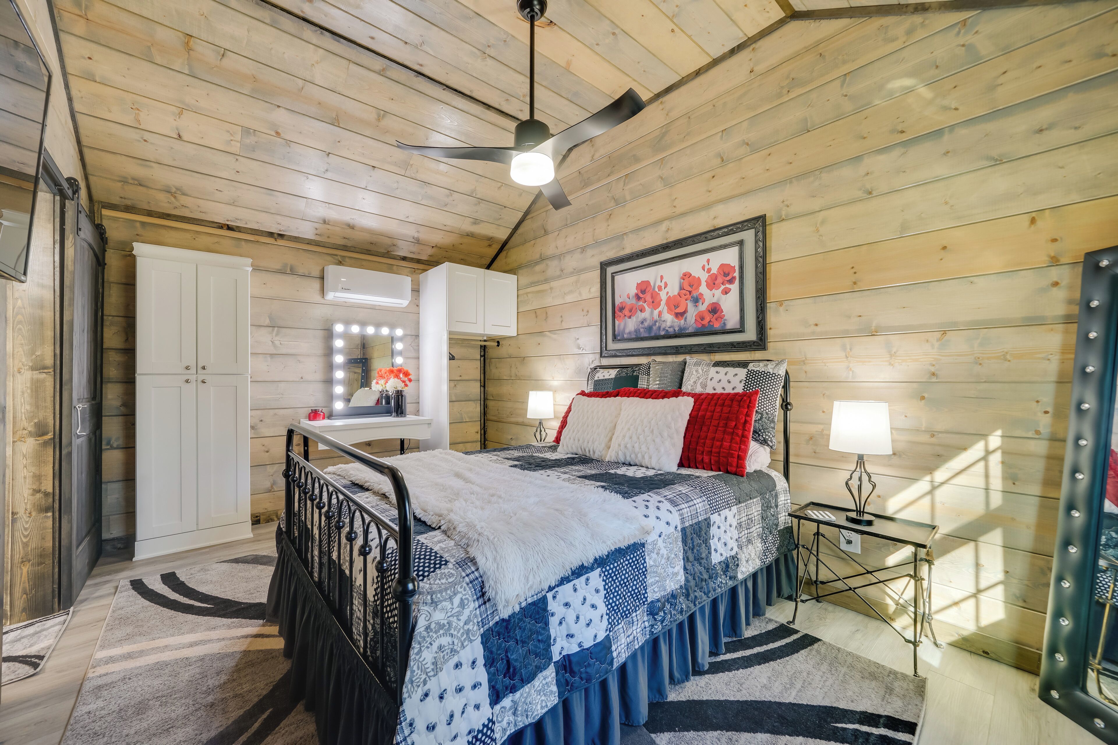 A cozy bedroom with a wooden ceiling, a bed with a quilt, a bedside table with a lamp, and a mirror above the bed.