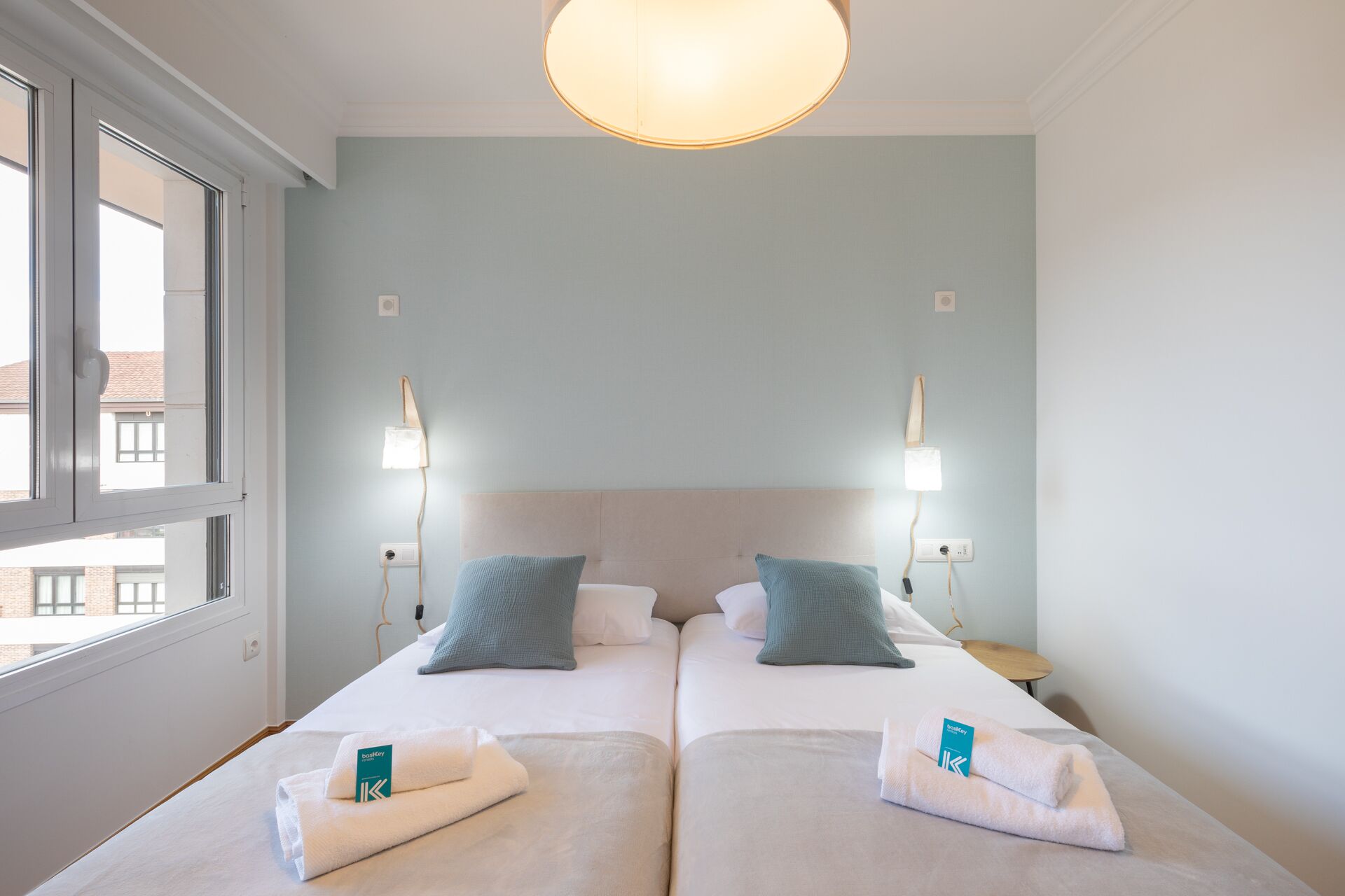Een tweepersoonskamer met twee bedden, elk met een kussen en een handdoek, een plafondlamp en een raam met uitzicht op gebouwen.