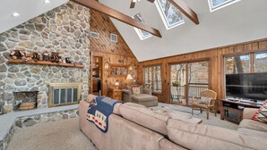 A cozy living room with a stone fireplace, wooden beams, and a large window overlooking a deck.