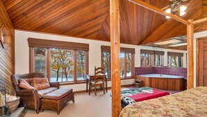 A spacious room with a large bed, a wooden ceiling, and a view of autumn trees through large windows.