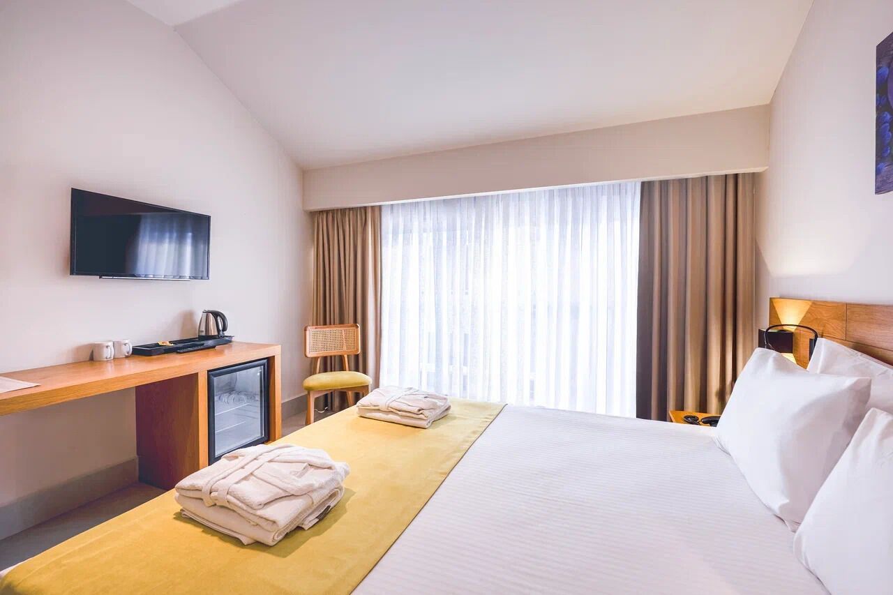 Una habitación de hotel con cama, escritorio con televisor, una silla y ventana con cortinas.