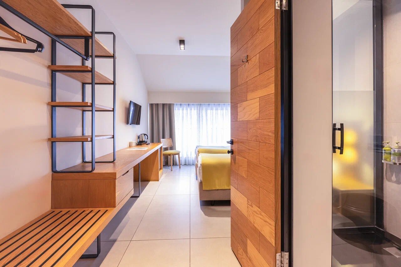 Una habitación de hotel moderna con un banco de madera, un escritorio, una cama con manta amarilla, un televisor montado en la pared y un box de ducha de vidrio.