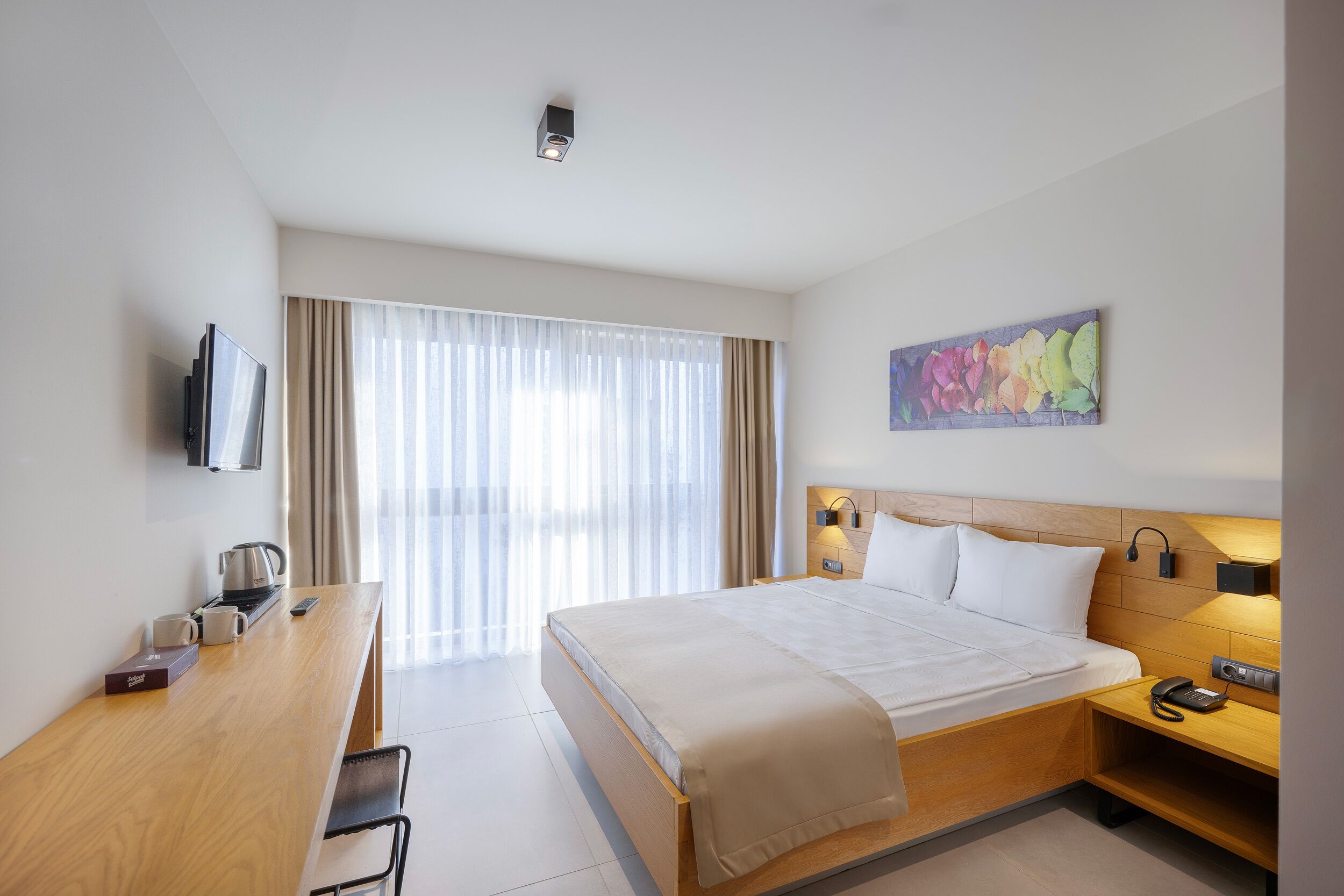 A hotel room with a bed, bedside table, lamp, TV, and a window with curtains.