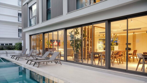 Un edificio moderno con piscina, sillas de descanso y un comedor visibles a través de amplios ventanales.