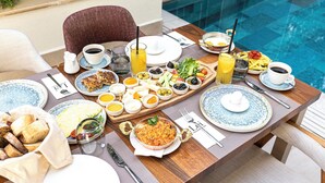 Una mesa de comedor con diversos alimentos para el desayuno, como huevos, tostadas y bebidas, cerca de una piscina.