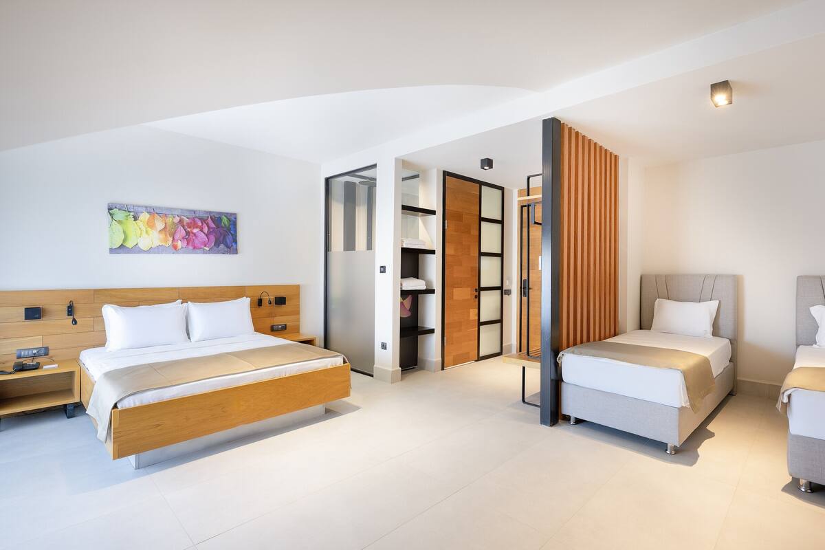 A modern hotel room with a large bed, a smaller bed, and a desk. There is a wardrobe with sliding doors and a shelf unit.
