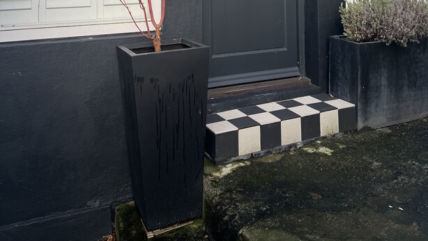Une porte à cadre noir, précédée d’un petit seuil, une enseigne au-dessus de la porte et un motif à carreaux sur le sol.