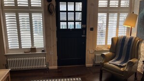 Une pièce avec une porte en bois foncé, des stores blancs, un radiateur, un tapis à motifs, un fauteuil en cuir avec un plaid à rayures et une lampe.