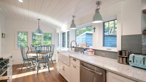 A kitchen with white cabinets, a stainless steel dishwasher, a blue toaster, and a window with a view of greenery. - Charming Cottage, Private Lake MI Beach (West Olive)