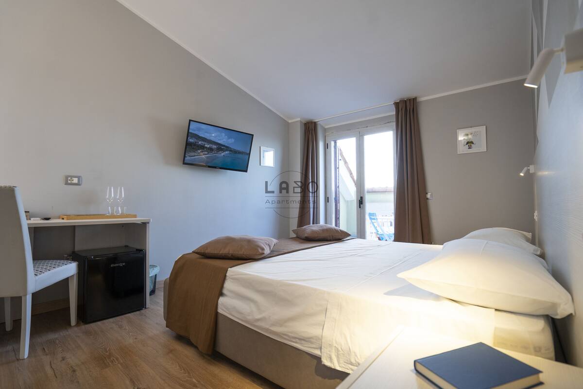 A hotel room with a bed, a desk, a chair, a small refrigerator, a TV mounted on the wall, and a window with curtains.
