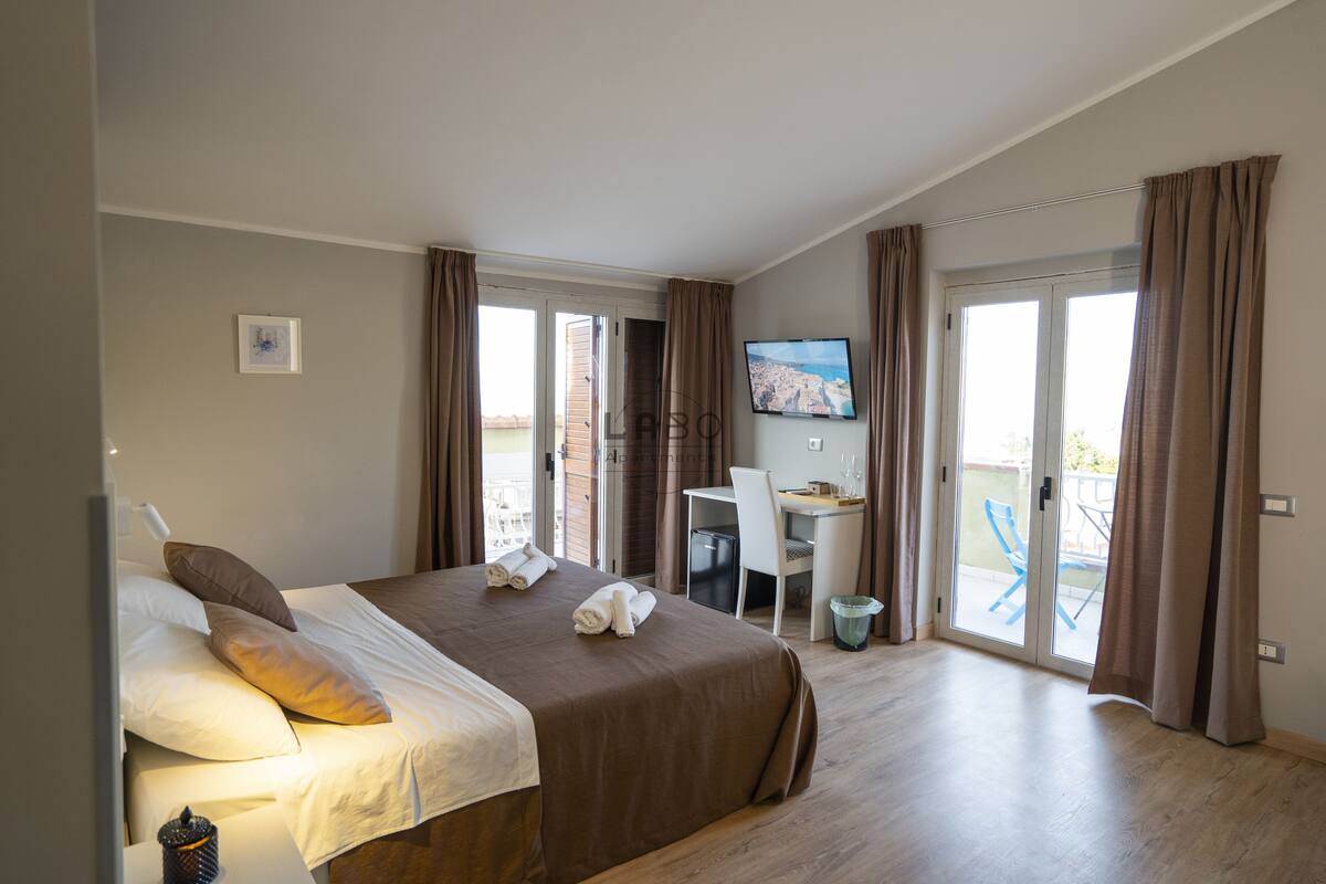 A hotel room with a bed, a desk, a chair, a TV, and a balcony with a chair.
