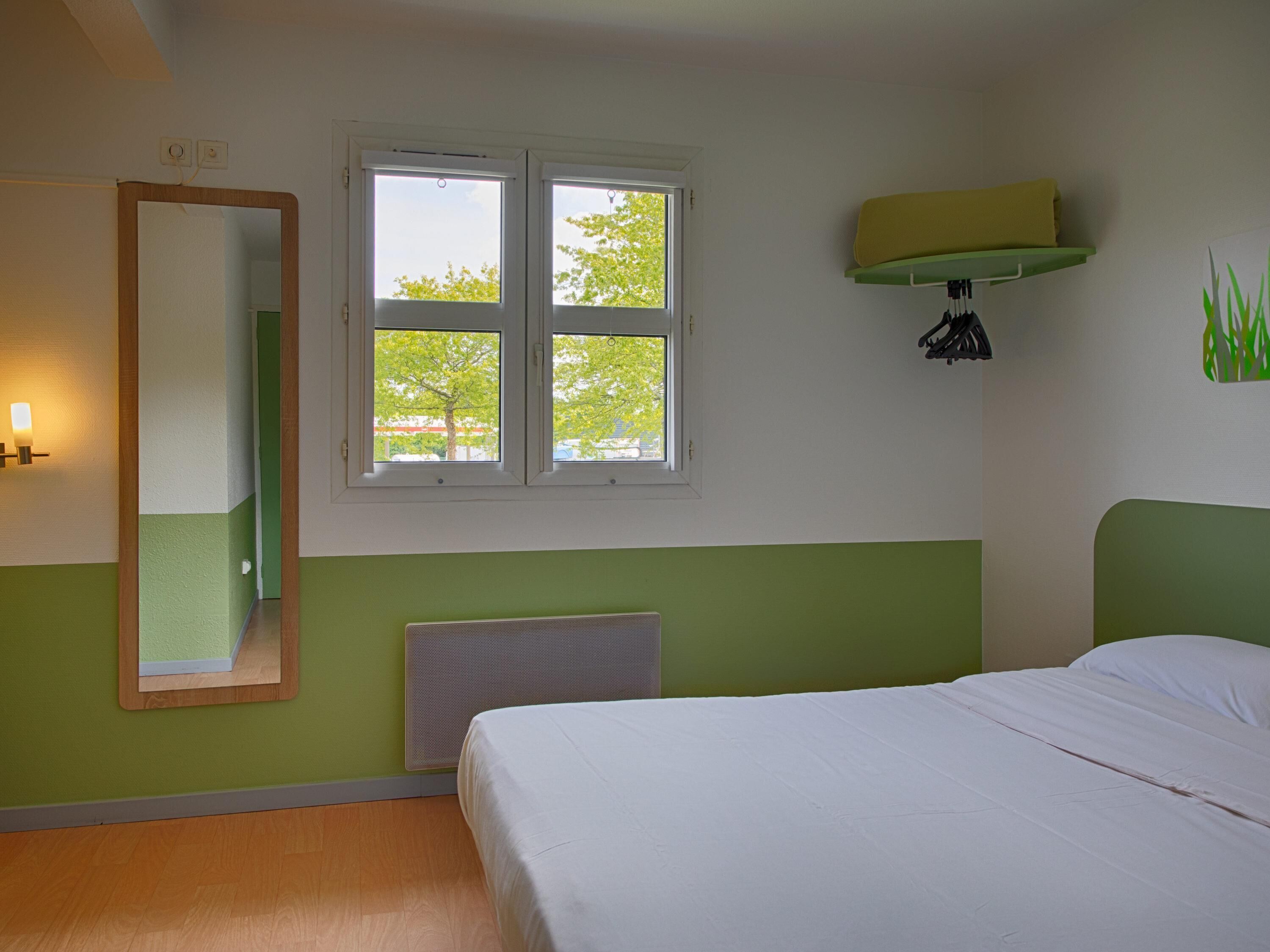 A bedroom with a bed, a window with a view of trees, a wall-mounted heater, and a mirror.