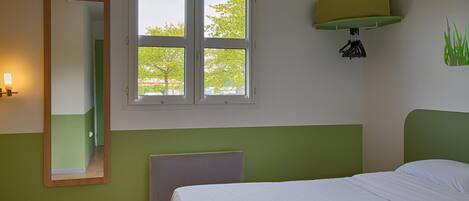 A bedroom with a bed, a window with a view of trees, a wall-mounted heater, and a mirror.