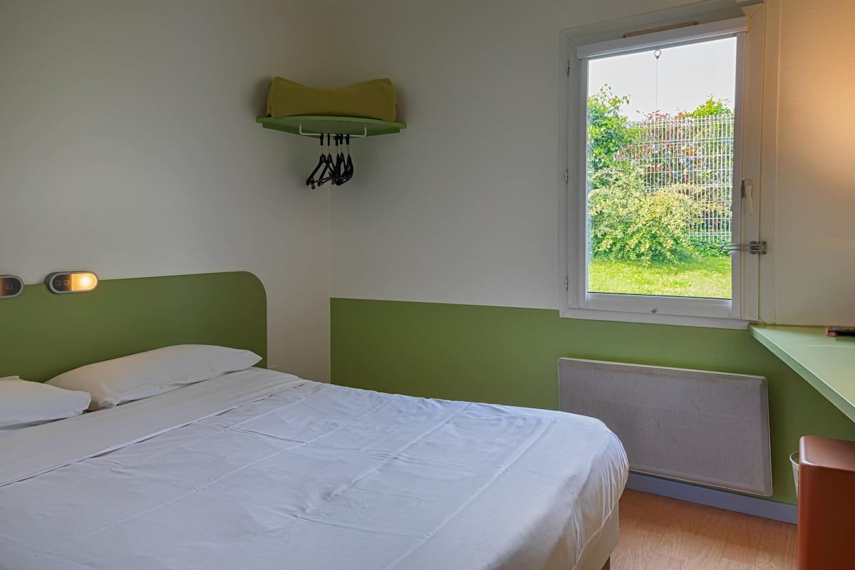 A bedroom with a bed, a window, a radiator, and a wall-mounted shelf with clothes.
