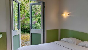 Un dormitorio con cama, pared verde decorativa, ventana con vistas a árboles y flores, y una lámpara fijada a la pared.
