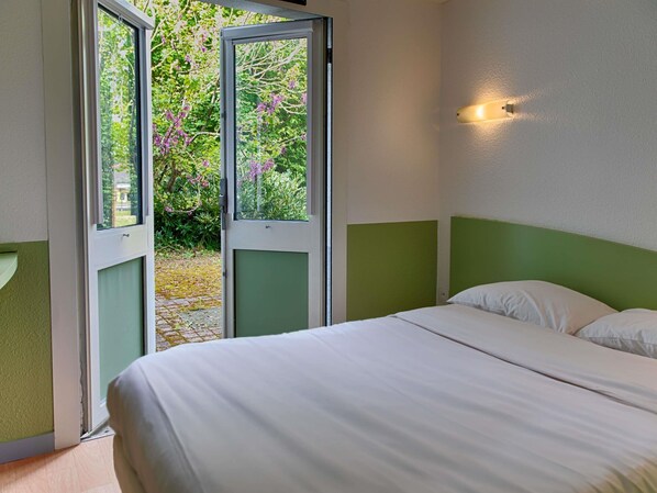 A bedroom with a bed, a green accent wall, a window with a view of trees and flowers, and a wall-mounted light.