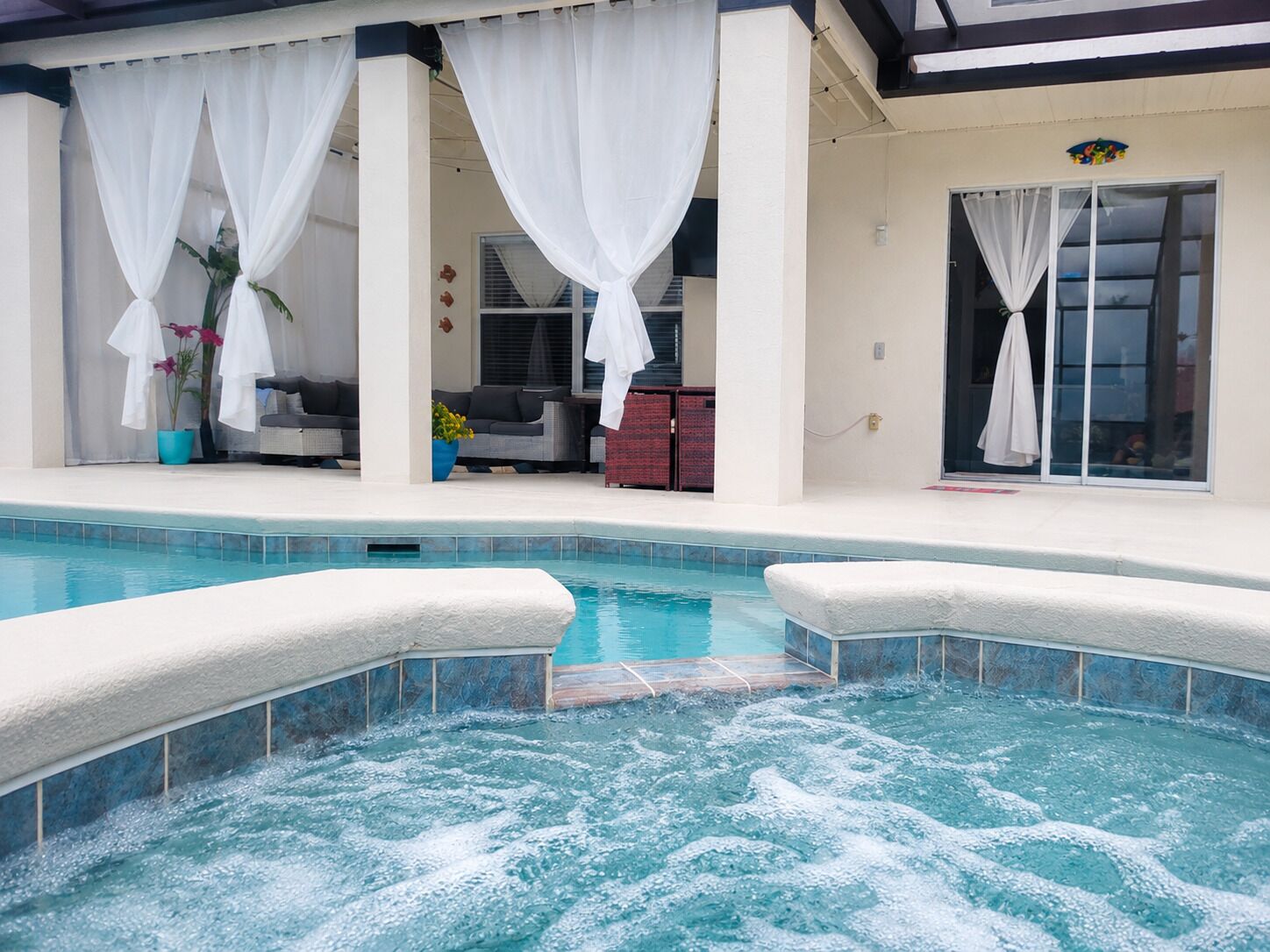 A swimming pool with a hot tub area, a covered seating area with a sofa and chairs, and a glass door entrance.