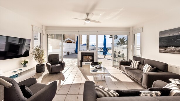 A modern living room with a flat-screen TV, comfortable seating, a fireplace, and large windows offering a view of the outdoors. - Faros Seaside Villa Larnaca (Pervolia)